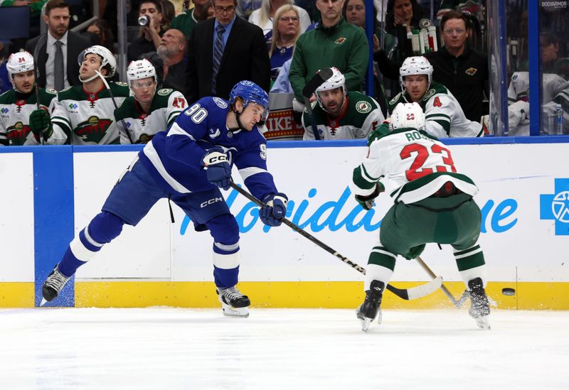 Oct 24, 2024; Tampa, Florida, USA; Tampa Bay Lightning defenseman J.J. Moser (90) shoots as Minnesota Wild center Marco Rossi (23) defends during the first period at Amalie Arena. Mandatory Credit: Kim Klement Neitzel-Imagn Images