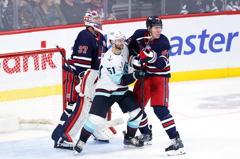 Oct 23, 2025; Winnipeg, Manitoba, CAN; Seattle Kraken center Shane Wright (51) and Winnipeg Jets defenseman Logan Stanley (64) wait for a incoming shot in front of Winnipeg Jets goaltender Connor Hellebuyck (37) in the second period at Canada Life Centre. Mandatory Credit: James Carey Lauder-Imagn Images