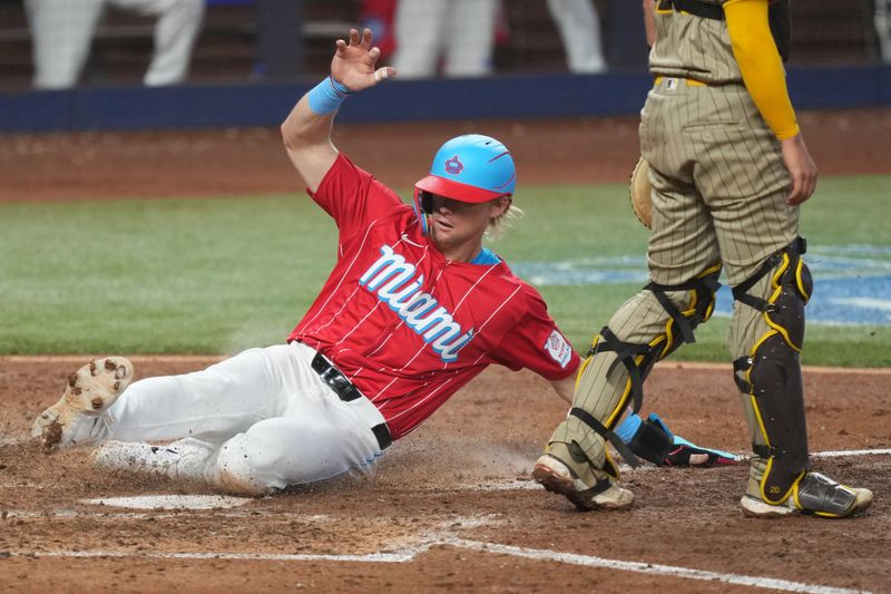 Aug 10, 2024; Miami, Florida, USA;  Miami Marlins left fielder Kyle Stowers (28) slides into home plate safely in the fifth inning as San Diego Padres catcher Kyle Higashioka (20) looks on at loanDepot Park. Mandatory Credit: Jim Rassol-USA TODAY Sports